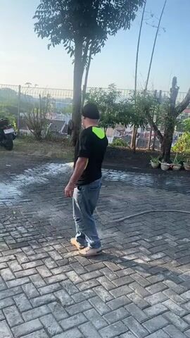 A person wearing a black shirt with a neon green collar and sleeves, blue jeans, and light-colored slip-on shoes stands in a courtyard. They are holding a hose and spraying water from it. The courtyard is paved with rectangular bricks. In the background, there is a tree, some other vegetation, potted plants, and a building. The sky is clear and blue, suggesting it is daytime. A motorcycle is parked to the left of the frame.