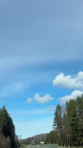 The video shows a highway scene. A red semi-truck is driving on the road, followed by a white truck. The sky is blue with scattered white clouds. Trees line both sides of the road.
