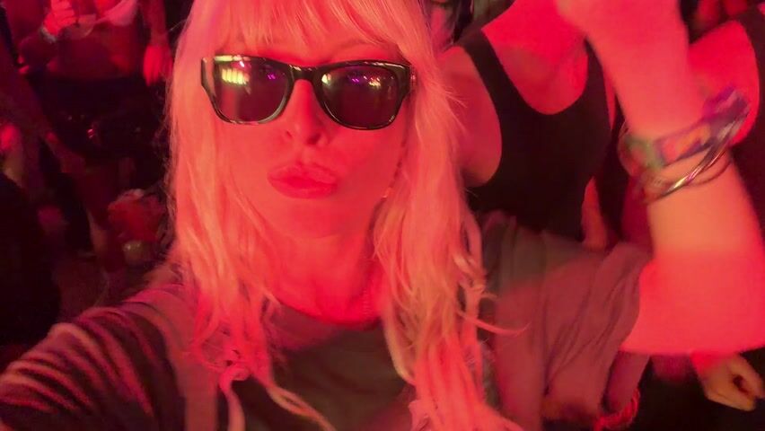 The video captures a blonde-haired woman, wearing sunglasses and a dark top, taking a selfie-style recording at a lively event. Her hair features a fringe, and she has several bracelets on her left wrist. She looks directly at the camera with a subtle pout or slight smile, conveying a sense of confidence and enjoyment.

The setting is a densely crowded indoor or tented venue, bathed in vibrant red artificial lighting. This lighting creates a dynamic and energetic atmosphere, illuminating the faces and figures of the attendees. In the background, numerous other people are visible, mostly out of focus, moving and dancing to the pulsating music, indicating a concert, club, or festival environment. The woman herself subtly sways and moves her head, appearing to be engaged with the music and the surrounding energy.

The heavy crowd density suggests a popular event. The artificial lighting throughout the scene indicates that it is likely nighttime. Given the location context of Indio, United States of America, the scene is highly suggestive of a major music festival such as Coachella, known for its vibrant nighttime experiences and distinctive lighting designs. No discernible text is visible on clothing or signs within the frame. The overall mood is one of exhilaration and celebration.