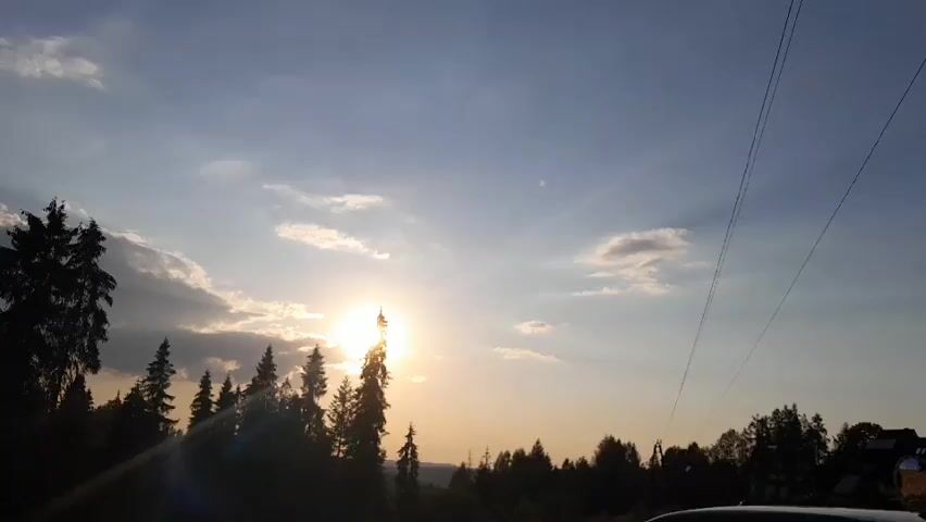 The video captures a sunset over a forested landscape with a clear sky and scattered clouds. The sun is visible in the center of the frame, partially obscured by trees and clouds, casting a warm glow. Silhouettes of evergreen trees line the left side of the frame, with more trees and a distant building visible on the right. Two power lines stretch diagonally across the upper right portion of the screen. The overall scene suggests an outdoor, possibly rural, environment during the golden hour.