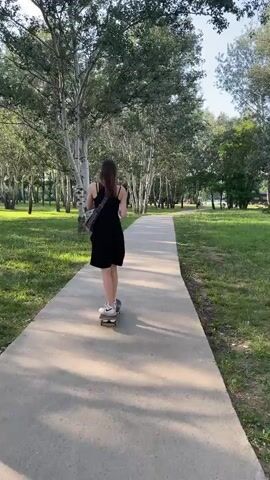A young person in a black dress is skateboarding on a paved path in a park. The person is facing away from the camera and is holding a bottle in their right hand. There are trees on either side of the path, and the sun is shining, casting shadows on the ground. The person is moving forward on the skateboard.