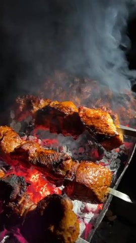 The video shows skewers of meat being grilled over glowing red coals at night. Smoke rises from the cooking meat, which appears to be marinated and is developing a charred crust. The camera is positioned close to the grill, focusing on the food and the flames. The background is dark, suggesting an outdoor setting at night.