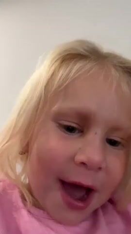 A young child with blonde hair and blue eyes is filmed from a close-up perspective. The child is wearing a pink shirt and appears to be laughing or making excited noises. The background is out of focus.
