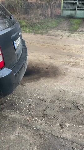 The video shows the rear of a dark-colored Jeep, with the license plate "BP 8143 CM." The vehicle is parked on a dirt road, which appears muddy. Thick black smoke is emitted from the exhaust pipe. The background features a blurred view of foliage, a house with a green fence, and an overcast sky.  The camera appears stationary, focusing primarily on the exhaust and surrounding ground.