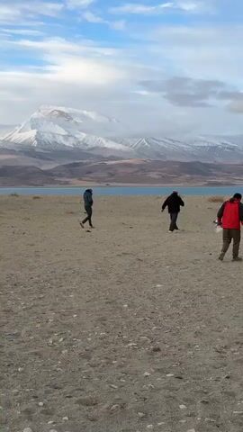 The video shows four people walking toward the edge of a lake. They are walking on a sandy, pebbly beach. A large mountain range is visible in the background, with snow-capped peaks partially obscured by clouds. The sky is blue with scattered clouds. One of the people is carrying a blue bucket, while another is carrying a white bag. They are walking in the same direction, but spread out across the beach.