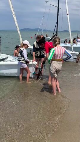 Four young people are standing in shallow water near a beach, gathered around the stern of a small sailboat. The boat is white with a mast and sail visible. The people are all wearing life jackets over their clothing. Two individuals in the foreground are looking down at something near the boat's rudder or keel, which appears to be a piece of equipment or possibly debris. One person is wearing a red shirt and tan shorts, another a striped long-sleeved shirt and tan shorts, and a third a white long-sleeved shirt and white shorts with a pink cap. The fourth person is wearing a dark life jacket over a dark shirt and shorts.

In the background, more sailboats are visible, some with people on board, suggesting a sailing event or school. The water is calm and clear, with gentle waves lapping at the shore. The scene appears to be taking place during the daytime, under a partly cloudy sky. The location is a beach with a sandy shore leading into the water. There are some distant landforms visible across the water, likely hills or mountains. The overall atmosphere seems to be one of activity and possibly instruction or teamwork related to sailing. No text is visible in the media.