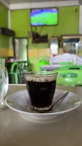 A medium shot captures a glass of dark coffee with foam on top, sitting on a white saucer with a spoon. The background is blurred, showing a room with green walls. A television screen displaying a soccer game is mounted on the wall. The environment appears to be a casual establishment like a cafe or restaurant, with tables and chairs visible. The lighting is natural and diffused.