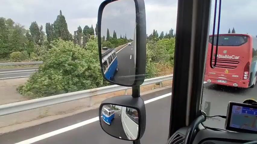 The video appears to be filmed from the driver's perspective inside a vehicle, possibly a truck or bus, on a highway. The view is directed forward and slightly to the side. In the foreground, the dashboard and side mirrors of the vehicle are visible. One side mirror shows the highway ahead, with a white line marking the lane.

The main subjects in the scene are two vehicles on the highway. Ahead of the recording vehicle, a blue and white vintage Volkswagen van (likely a Type 2 "Kombi") is driving in the right lane. Behind the van, further down the highway, is a large red bus. The red bus has "BlaBlaCar" written on its rear in white lettering, indicating it is a ride-sharing service. The highway is a multi-lane road with a metal guardrail on the left side.

The setting is a highway surrounded by lush green trees and vegetation, with some cypress trees visible, suggesting a Mediterranean or southern European landscape. The weather appears overcast, with a grey sky. The time of day seems to be daytime, likely afternoon, given the light conditions.

The activity taking place is the vehicles traveling on the highway. The vintage van is in front of the recording vehicle, and the red bus is further ahead of the van. The overall impression is a snapshot of a road trip in what appears to be a scenic but somewhat cloudy environment.

Visible text includes "BlaBlaCar" on the back of the red bus. There's also a signpost visible in the distance on the right side of the highway. Although the text on the sign is blurry, it appears to contain route information or place names, possibly indicating "GRANDS CROMOS" and "800m" followed by a number that could be a road number. A GPS device is also mounted on the dashboard of the recording vehicle.