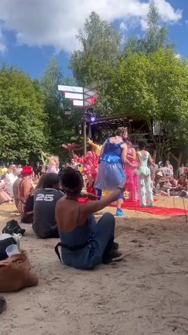 The video shows an outdoor event during daytime. In the foreground, a person wearing a blue jumpsuit and a headband is sitting on the sand, facing away from the camera. They are looking towards a stage in the background where several individuals are dancing. The stage is adorned with lights and a red carpet. The dancers are wearing colorful costumes, including a blue dress, a yellow dress, and pink dresses. The event appears to be a performance or festival, with an audience sitting on the sand in the midground. The environment is a sandy area surrounded by trees and a blue sky with clouds. The person in the foreground is gesturing with their right hand. Another person, wearing a dark jacket and pants, is walking past the foreground person.