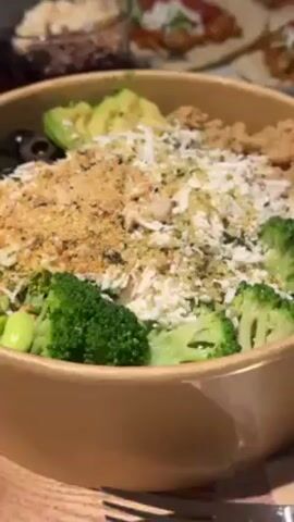 The video shows a variety of healthy food options laid out on a table. In the foreground, there's a tan bowl filled with a salad containing broccoli, avocado, cheese, and other toppings. Behind the salad, there's a small glass bowl containing a dark purple base mixed with granola, chocolate nibs, and coconut flakes, accompanied by a wooden spoon. Next to the glass bowl, a plate holds several small tacos topped with lettuce, salsa, cheese, and other fillings. A small glass of sauce sits nearby. The table has a gray surface. The lighting is dim, giving the scene a casual feel. The camera pans slowly across the food, focusing on each item for a moment.