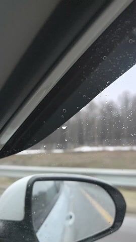 Here is a brief description of the video:

The video appears to be taken from inside a car, likely the driver's perspective. Rain or snow is falling outside, visible on the window. The car's side mirror and a blurred roadside are visible in the background. Music is playing.