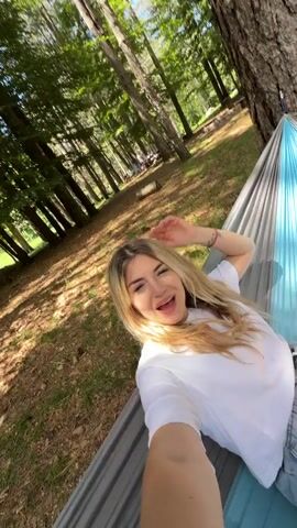 A woman with long blonde hair and a white t-shirt and blue denim shorts lies in a hammock in a forest. She smiles and then shows her legs in white socks, and then shows herself smiling in the hammock again. A picnic blanket and shoes are on the ground in the background.