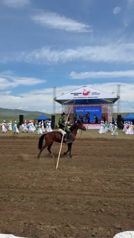 A man in traditional Mongolian clothing, a dark hat and green garment, rides a brown horse. He holds a long, thin pole extending from the horse's saddle forward and backward, suggesting a traditional game or performance. The horse is walking in a dirt arena. In the background, a stage is set up with a large banner displaying text and a swan logo. Several people in white and colorful traditional attire are gathered on and around the stage, possibly performing or watching. Two men with cameras are visible to the right of the stage, filming the event. The environment appears to be an outdoor field with rolling hills and a bright blue sky with scattered clouds. The overall scene depicts a cultural event likely celebrating traditional Mongolian sports or festivities.
