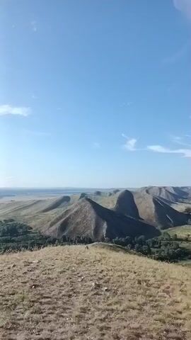 The video shows a panoramic view of rolling hills under a bright blue sky with scattered clouds. In the foreground, there are the tops of trees and the undulations of the landscape. Further back, the hills become more pronounced with dry grass and some trees. In the distance, a body of water or flat land can be seen. The camera pans and tilts, revealing a white church with a golden dome and a cross on top, situated on a hilltop. The church is viewed from multiple angles, including upside down at one point, suggesting a dynamic camera movement, possibly with a drone.