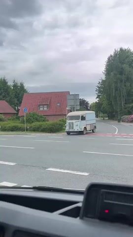 The video shows a vintage light blue and beige Citroen van driving through an intersection. The van has a white roof and a license plate. It is driving on a paved road with white lines. On the side of the road is a red brick building with a matching red tiled roof, some green hedges, and a street sign. The sky is overcast and gray. There are trees in the background. A dark gray modern SUV drives past the camera's viewpoint, briefly obscuring the Citroen van. The video appears to be shot from inside a vehicle.