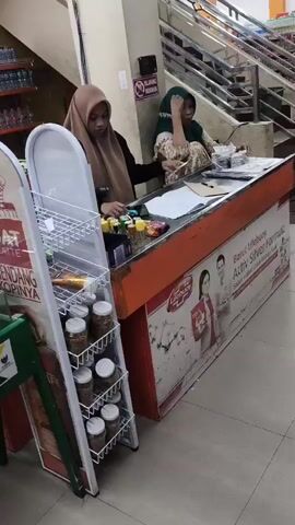 A video shows two women behind a checkout counter in what appears to be a store. The woman on the left, wearing a brown hijab, stands behind the counter and looks towards the right. The woman on the right, wearing a green hijab, is seated and is engaged in activity behind the counter, possibly handling money or items. In front of the counter, a customer, a woman wearing a plaid shirt and dark pants, approaches the counter. She is holding her phone in her right hand and looking at it. A display rack with jars is to the left of the counter. Stairs are visible in the background.