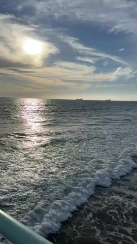 The video shows a view of the ocean from a slightly elevated perspective, likely a pier or a boat. The sun is visible in the upper left quadrant, partially obscured by clouds, casting a shimmering reflection on the water's surface. Several ships are visible on the horizon. The water is choppy with visible waves and foam breaking near the foreground. A portion of a light-colored railing is visible in the bottom left corner. The sky is blue with scattered clouds.