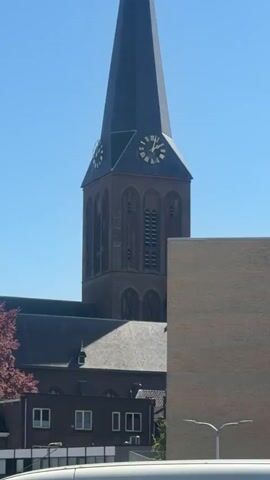 The video shows a tall, brick church tower with a clock on its face against a clear blue sky. The camera pans out to show the church building and some nearby buildings, including an apartment complex. The shot ends with a view from a parking lot, with cars in the foreground and the church steeple visible in the background. The environment appears to be urban.