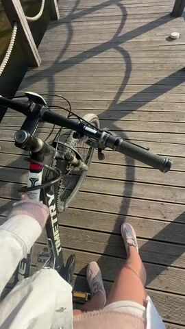 The video captures a sunny outdoor scene from a first-person perspective on a wooden pier. It begins with a close-up view of a person's legs, wearing light pink sneakers and shorts, standing next to a black bicycle. The bicycle frame prominently displays "TIMETRY" in white lettering, and "Totem" is visible on the front fork. Strong sunlight casts distinct, long shadows of the pier's railings and the bicycle across the wooden planks.

The camera then pans up and across the scene, revealing the wooden pier extending into a large, calm body of water, likely a lake or wide river. The pier features dark wooden posts connected by thick rope railings. The sky is bright blue with scattered white clouds. The sun is directly overhead, creating a brilliant glare and reflections on the water's surface. In the distance, the shoreline reveals a mix of buildings, possibly residential complexes or industrial structures, along with several tall cranes or towers, suggesting an urban or developing landscape. The setting is consistent with a waterfront recreational area, possibly in Myakinino, Russia, enjoying a clear and bright day.