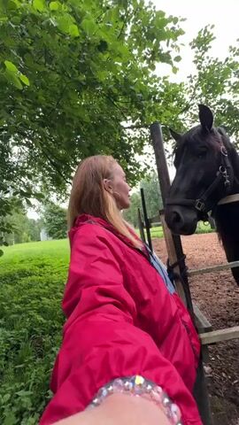 A woman with blonde hair, wearing a bright red jacket over a blue top, stands in a lush green outdoor setting. She is smiling and looking towards the camera, holding it out to capture a selfie. To her right, a black horse with a bridle stands behind a wooden fence. The horse is looking towards the woman. The background shows more greenery, trees, and a cloudy sky, suggesting an overcast day. The scene appears to be a farm or a rural area, with a wooden fence and what looks like a horse enclosure. The woman has a bracelet on her wrist with colorful beads. The overall mood is cheerful and friendly, with a focus on the interaction between the woman and the horse. The setting evokes a sense of nature and connection with animals. There is no visible text in the media file.