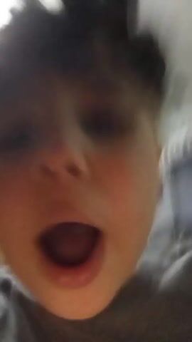 Okay, here is a concise description of the video:

The video shows a young boy, close-up, speaking directly to the camera. He has dark curly hair, light skin, and is wearing a gray t-shirt. He is looking at the camera and his mouth is open as if he is talking or about to speak. The background is blurry but shows hints of an interior room with a light-colored wall and possible decorations. The camera is shaky.