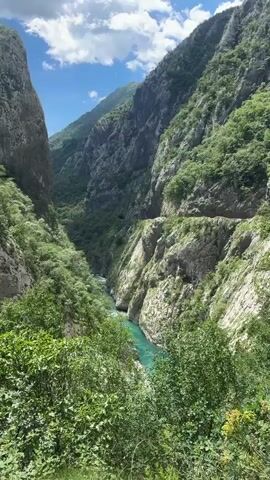 The video shows a picturesque view of a deep river canyon surrounded by towering mountains with a bright blue sky and scattered white clouds overhead. The river below is turquoise in color and winds through the canyon floor, flanked by lush green vegetation. The canyon walls are steep and rocky, with varying textures and patches of greenery clinging to their surfaces. The camera pans upwards to show the full height of the mountains and the sky above. A tunnel entrance is visible in the distance on the canyon wall.