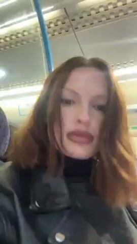 A woman with brown hair and dark lipstick applies lip balm on a train. She is wearing a black turtleneck sweater and a black leather jacket. The train car has purple seats and a white ceiling with fluorescent lights.