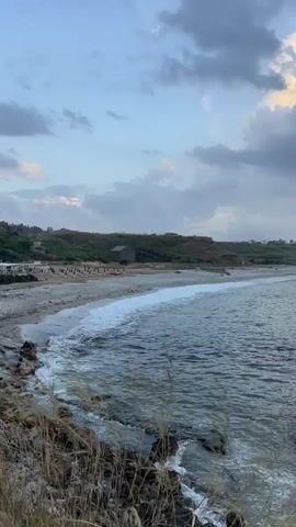 The video shows a wide shot of a rocky beach and the ocean. The sky is cloudy, and the sun is setting. Waves crash against the shore. In the distance, there are some buildings and trees. The overall impression is one of a peaceful and serene setting.