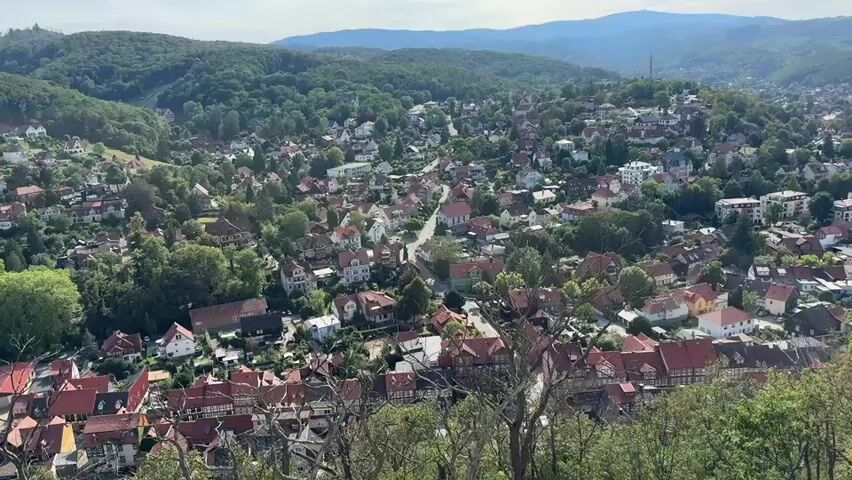 This video provides a panoramic view of a densely populated town situated on a hillside. The town features a mix of traditional and modern architecture, with many houses displaying red-tiled roofs, characteristic of European villages. Lush green trees and foliage are interspersed throughout the residential areas, creating a natural and scenic environment. In the background, rolling hills covered in dense forests suggest a rural or semi-rural setting. The sky appears to be a hazy blue, indicating a possibly overcast or misty day. There are no people or animals visible in the video, and the focus is solely on the landscape and the built environment. The camera seems to be positioned at a high vantage point, looking down and across the town.
