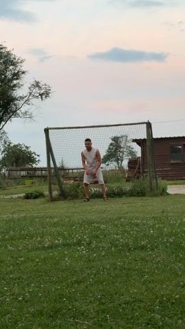 A man in a white tank top and light-colored shorts stands in front of a soccer goal in a grassy field. He is holding a red object, possibly a ball, in his right hand. The sky above is a soft pink and blue, suggesting it is either sunrise or sunset. Trees and a wooden building are visible in the background, with a fence and some gardening supplies near the goal. The man appears to be preparing to play soccer. He kicks the ball into the air and then watches it as it ascends. He then walks away from the goal. The setting appears to be a rural or semi-rural area, with a relaxed and casual atmosphere. There is no visible text in the media file.