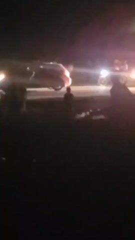 At night, the video shows a road scene. Several blurred figures stand on what appears to be a roadside area. Vehicles, including what seems to be a car and a motorcycle with multiple riders, are on the road, their headlights and taillights visible. A police car with flashing blue and red lights is also present. The overall visibility is poor due to the darkness.