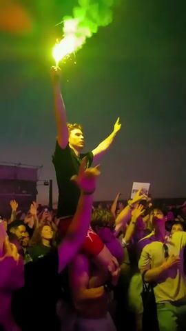 A low-angle shot captures a crowd at an outdoor event, likely a concert, under a dusky sky. In the foreground, several people are visible, their faces turned upwards. The central focus is a young man being carried on the shoulders of another individual. The man on the shoulders is holding a lit green flare high in the air, its bright smoke billowing upwards and casting a vibrant green glow on the scene. The crowd around him appears excited, with many people raising their hands in the air and smiling. The atmosphere is energetic and festive.