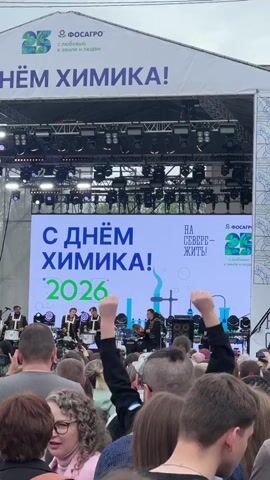 A crowd of people faces a stage with a large LED screen displaying the text "С ДНЁМ ХИМИКА! 2026" and "НА СЕВЕРЕ - ЖИТЬ!". A band is performing on stage; they are wearing black outfits and playing instruments including drums and guitars. The crowd is diverse in age and appearance, and some are raising their hands in excitement. The event appears to be sponsored by FOSAGRO, as their logo is prominently displayed on the stage banner. The environment suggests an outdoor celebration or festival.