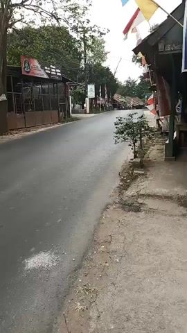 The video captures a street scene from a low angle, suggesting it was filmed from a moving vehicle or held by someone walking. On the left, there are several buildings with shaded awnings, some featuring signage with red lettering and a circular logo. A road stretches ahead, appearing slightly damp. On the right side of the road, there is a sidewalk with dirt and some vegetation, including small trees. Further down the road, on the right, are more structures, one of which has a large poster depicting two soccer players, one of whom resembles Lionel Messi. Flags, possibly national or decorative, are visible on poles in front of some buildings. A motorcycle with two riders is seen approaching and passing by in the middle of the road. The overall environment suggests a commercial or residential street in a tropical or subtropical region, possibly during a celebration or event given the flags and decorations.