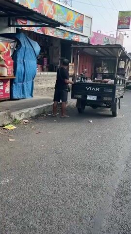 A man wearing a black t-shirt and black shorts is standing on an asphalt road next to a black truck with the brand VIAR. The truck has a canopy and appears to be a food cart, with various items displayed on its surface. The man is holding something in his hands, possibly food or money, and is facing the truck. To the left of the truck and man, there is a sidewalk with a store or market behind it. The store has a blue tarp covering some of its offerings and a colorful banner with the number 76. The environment appears to be an outdoor market or street vendor area, with some debris on the ground. The lighting suggests it is daytime, with some shadows present.