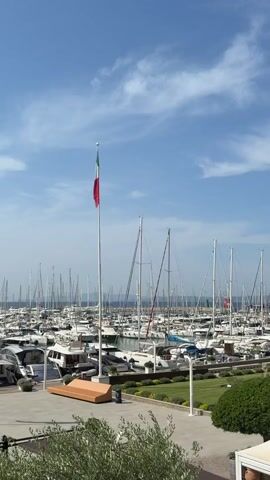 A medium shot captures a harbor filled with numerous sailboats and motorboats docked under a clear blue sky with scattered clouds. In the foreground, a tall flagpole stands to the left, flying the Italian flag. A wooden bench is positioned on a paved area in front of the flagpole, with a trash can nearby. A green lawn with hedges borders the paved area. The background shows a dense collection of boats, their masts creating a complex pattern against the horizon. The overall scene depicts a serene and well-maintained marina on a sunny day.
