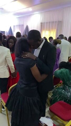The video shows a gathering of people in what appears to be a banquet hall or event space. In the initial shot, a man in a black suit embraces a woman in a black dress, with another couple holding hands nearby. The camera then moves to show a wider view of the room, revealing rows of red and gold chairs, and people dressed in various outfits milling about or taking their seats. The decor includes sheer white curtains and black decorative accents. The room is brightly lit.