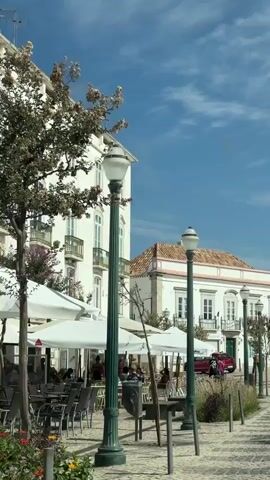 The video pans from left to right, showing a European-style town square on a sunny day. In the foreground, there are cobblestone paths and metal posts. To the left, there are outdoor cafe tables with white umbrellas and several people seated. Tall, dark green lampposts with white globes are spaced throughout the scene. Behind the cafe, a multi-story white building with numerous windows and balconies is visible. Further back, another white building with a terracotta roof and arched windows is present. Several cars are parked along the street. In the middle ground, a person walks away from the camera up a set of stairs. The background features more white buildings, trees, and a blue sky with scattered clouds. The overall impression is of a pleasant, well-maintained urban public space.