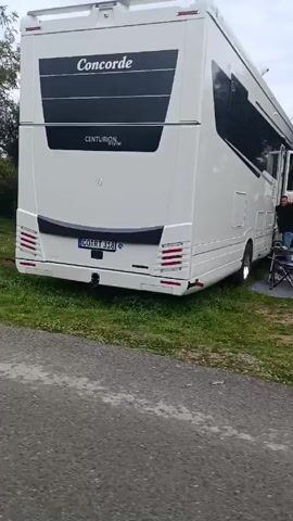 The video shows a motorhome parked on a grassy area with trees nearby. The motorhome is white and has "Concorde" and "CENTURION style" written on the back. The license plate reads "CO RT 318." The camera pans to the side, showing more of the grassy area with trees and a clothesline. The camera moves forward along a narrow asphalt road. Finally, the view is obscured by what appears to be a dirty, cracked window or windshield.