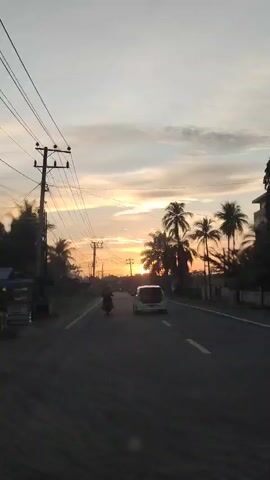 The video shows a view from inside a car driving down a road lined with palm trees, at sunset. The sky is a mix of yellow, orange, and grey. Overhead electrical wires are also visible. There is a car and two motorcycles further down the road ahead. The road has yellow lines dividing the lanes.