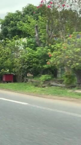 A motorcycle rider wearing a helmet and a tan uniform with red accents passes by on the right side of the road. The rider is on a black motorcycle. The environment appears to be a rural or suburban area with trees and buildings visible along the roadside. There are signs and some construction materials in the background. The perspective suggests the video is filmed from another vehicle moving in the same direction.