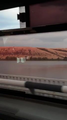 The video shows a view from a moving vehicle, looking out a window. The scene outside consists of a body of water and a large, reddish-brown hill with some vegetation at its base. The sky appears cloudy. The foreground shows the interior of the vehicle, with parts of the window frame and some horizontal bars visible. Reflections of the vehicle's interior are also visible on the window. The video captures a short duration of travel, with the landscape slowly moving past.