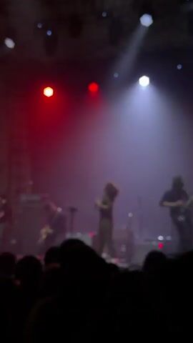 A band performs on a stage under a dynamic light show for a crowd of people. The band consists of three members: a female vocalist in a jumpsuit, and two guitarists in dark clothing. The lighting alternates between white spotlights and a wash of red light, highlighting the performers as they play. The silhouettes of the audience are visible in the foreground, with some individuals holding up phones. Musical equipment is visible on stage.