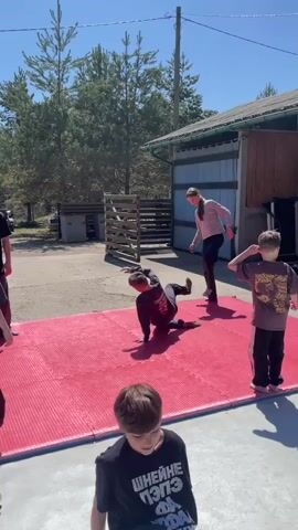 Outside, on a concrete patio beneath a canopy, a group of young people are practicing dance or martial arts moves. Some are on a large red mat, while others sit or kneel on the concrete floor, apparently taking a break.  Their shoes are piled in a corner.  The area is sunny, and the background shows a building, trees and an open area.  One person is wearing a striped shirt and loose pants, while the others are in dark clothing.  The general atmosphere seems relaxed and casual.