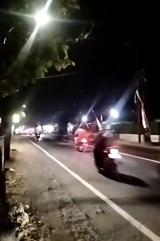 A group of motorcyclists are riding down a street at night. The street is illuminated by streetlights and the headlights of the motorcycles. Some of the motorcycles have flags attached to them. The motorcyclists are wearing dark clothing. The street is lined with trees and buildings. The pavement is wet, suggesting it may have recently rained. The motorcycles are moving at a moderate speed. The overall impression is of a nighttime motorcycle parade or procession.