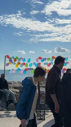 The scene is outdoors, featuring a stone breakwater and a sunny sky with scattered clouds. A long frame covered in colorful balloons is set up in front of a blue sea, where a large ship is visible in the distance. People are walking by, including a woman with long dark hair. A man is sitting on the rocks, and there's a black plastic crate nearby. Graffiti is visible on the rocks.