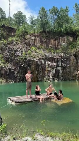 A group of four people, three women and one man, are on a raft in a clear blue lake. The man stands on the raft holding a phone and filming. The three women are sitting on the raft. One woman in a black swimsuit is in the middle of the raft. Another woman in a pink swimsuit is partially submerged in the water at the edge of the raft. A third woman in a black swimsuit is sitting next to the woman in the pink swimsuit. The raft is made of bamboo. In the background, there is a large rock cliff face with trees on top. The sky is blue with some clouds.