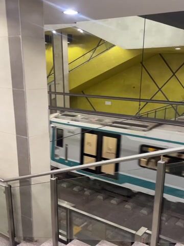 The video shows a modern metro station. A white train with a teal stripe is seen approaching and then stopping at a platform. The camera is positioned from an upper level, looking down at the tracks and the platform. In the foreground, there are stairs with glass and metal railings. A large, light-colored pillar is prominent on the left side of the frame initially, partially obscuring the train.

As the train slowly pulls into the station, more of the station's architecture becomes visible. The station features striking bright yellow walls with a grid-like pattern of black cables or bars running across them. Numerous light fixtures are mounted on the ceiling, illuminating the yellow walls. Escalators are visible in the background, leading to other levels of the station.

The train's destination display shows the word "ISIDOR". People can be seen inside the train cars through the windows, and some figures are visible on the platform as the train comes to a halt. The overall impression is that of a clean, well-lit, and architecturally distinct underground or elevated transit station. The sound of the train's brakes and movement is audible throughout the video.