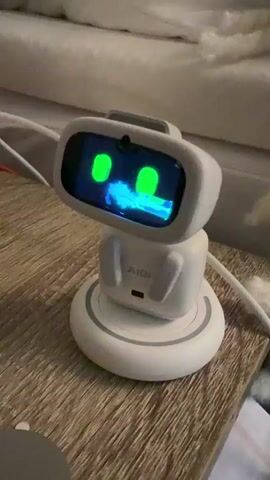 The video shows a small, white robot with a screen for a face. The robot is sitting on a wooden surface, possibly a bedside table. The screen displays a cartoon face with green eyes and a blue, possibly animated, mouth. The robot appears to be in a room with soft furnishings in the background, suggesting an indoor setting. White cables are visible around the robot, indicating it is likely plugged in or charging. The lighting is soft, and the overall impression is one of a quiet, domestic environment. The time of day is not specified. The robot's display is the central focus, showing a simple animated expression. The brand name "AIBI" is visible on the robot's torso. No people are visible in the frame. The setting suggests a bedroom or living space.