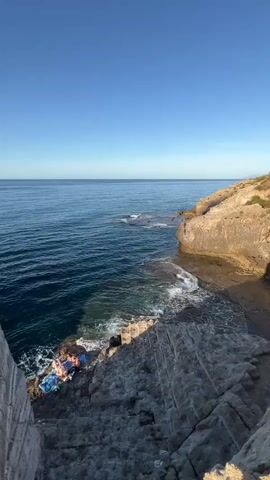 A coastal scene unfolds with a clear blue sky overhead and a vast expanse of blue ocean stretching to the horizon. On the right, light brown cliffs rise from the water, while in the foreground and to the left, grey, layered rock formations descend towards the sea. Gentle waves crash against the rocky coastline. Two individuals are observed on the lower grey rock area, near the water's edge, appearing to relax on towels. The camera pans slightly down and then zooms in on the ocean and the two individuals.